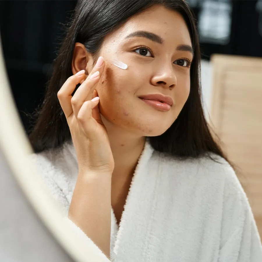 photo of young asian woman with brunette hair applying acne treatment cream and looking at mirror